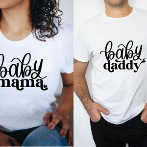 May include: Two white t-shirts with black text. One shirt says "baby mama" and the other says "baby daddy".