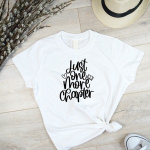 May include: White t-shirt with black text that reads "Just one more chapter" with a heart and a book icon.