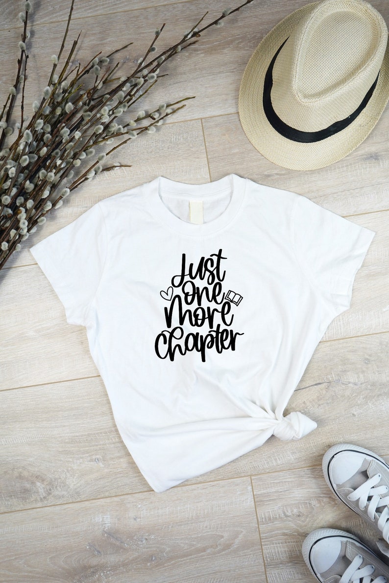 May include: White t-shirt with black text that reads "Just one more chapter" with a heart and a book icon.