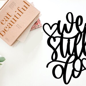 May include: Black metal sign with the words "we still do" in cursive script. The sign is decorated with three small hearts.