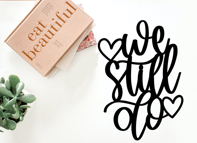 May include: Black metal sign with the words "we still do" in cursive script. The sign is decorated with three small hearts.