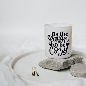 Puede incluir: Vela blanca con una inscripci&oacute;n negra que dice "'tis the season to be cozy" con dos peque&ntilde;os corazones a cada lado de la palabra "to". La vela est&aacute; sentada sobre dos rocas grises sobre una superficie blanca.