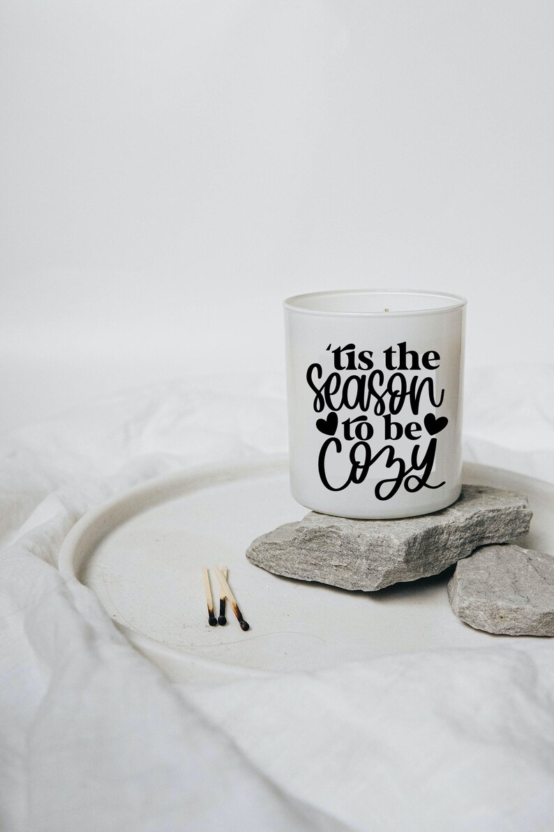 Puede incluir: Vela blanca con una inscripci&oacute;n negra que dice "'tis the season to be cozy" con dos peque&ntilde;os corazones a cada lado de la palabra "to". La vela est&aacute; sentada sobre dos rocas grises sobre una superficie blanca.
