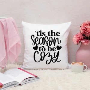 Puede incluir: Almohada blanca con texto negro que dice 'Tis the season to be cozy' con dos peque&ntilde;os corazones negros a cada lado de la palabra 'be'.