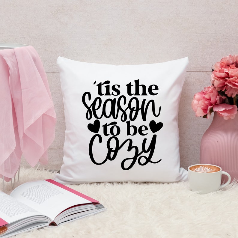 Puede incluir: Almohada blanca con texto negro que dice 'Tis the season to be cozy' con dos peque&ntilde;os corazones negros a cada lado de la palabra 'be'.