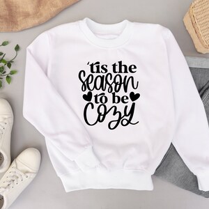 Puede incluir: Sudadera blanca con texto negro que dice "'Tis the season to be cozy" con dos peque&ntilde;os corazones a cada lado de la palabra "be".