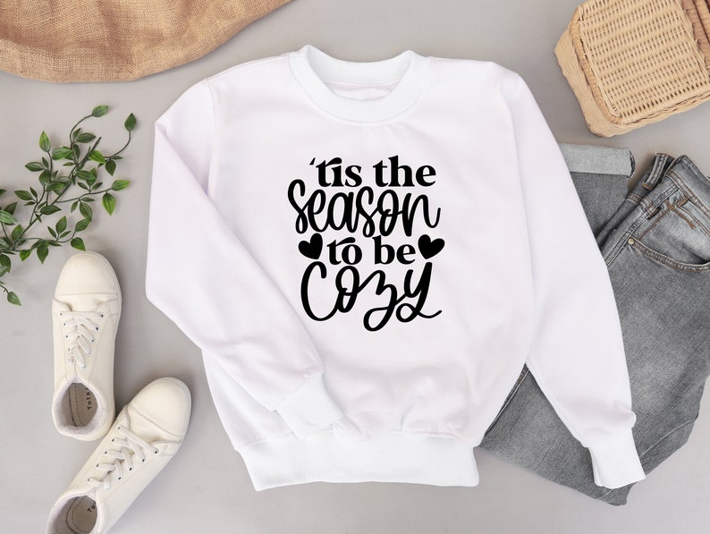 Puede incluir: Sudadera blanca con texto negro que dice "'Tis the season to be cozy" con dos peque&ntilde;os corazones a cada lado de la palabra "be".
