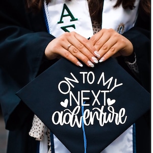 May include: A black graduation cap with a white tassel and the text "On to my next adventure" written in white cursive lettering with two small hearts on either side of the word "next".