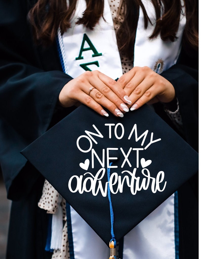 May include: A black graduation cap with a white tassel and the text "On to my next adventure" written in white cursive lettering with two small hearts on either side of the word "next".
