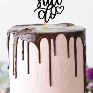 May include: A pink and chocolate cake with a black cake topper that says "We Still Do" with hearts on either side.