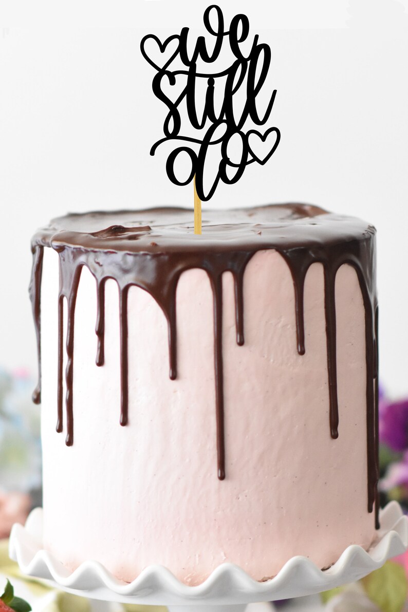 May include: A pink and chocolate cake with a black cake topper that says "We Still Do" with hearts on either side.