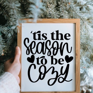 Puede incluir: Un cartel enmarcado con el texto 'Tis the season to be cozy' en letra cursiva negra con dos peque&ntilde;os corazones a cada lado de la palabra 'be'. El cartel se sostiene frente a un &aacute;rbol de Navidad verde.