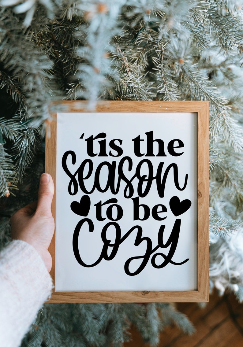 Puede incluir: Un cartel enmarcado con el texto 'Tis the season to be cozy' en letra cursiva negra con dos peque&ntilde;os corazones a cada lado de la palabra 'be'. El cartel se sostiene frente a un &aacute;rbol de Navidad verde.