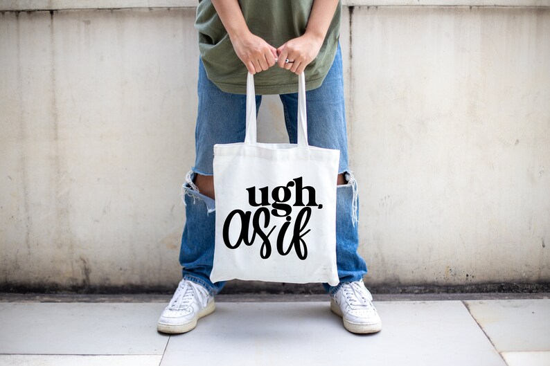 May include: A white canvas tote bag with the text "ugh, as if" printed in black. The bag is being held by a person wearing blue jeans and white sneakers.