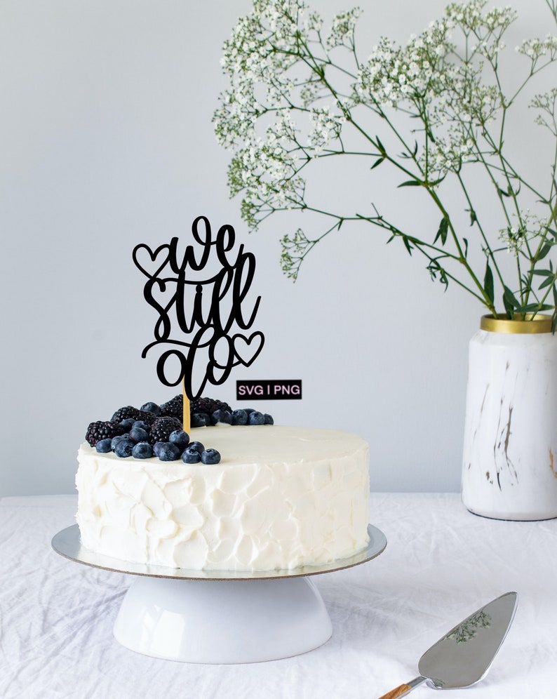 May include: A white cake with a black topper that says "we still do" with hearts. The cake is decorated with blueberries and blackberries. The cake is on a white cake stand. There is a vase of white flowers in the background.