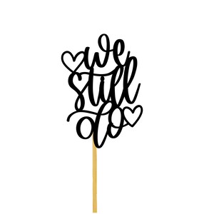 May include: Black cake topper with the text "we still do" and two hearts. The topper is on a wooden stick.