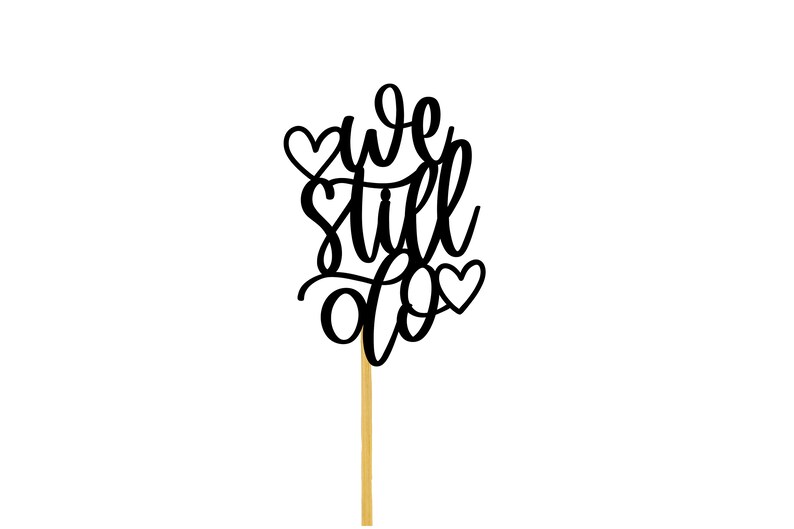 May include: Black cake topper with the text "we still do" and two hearts. The topper is on a wooden stick.