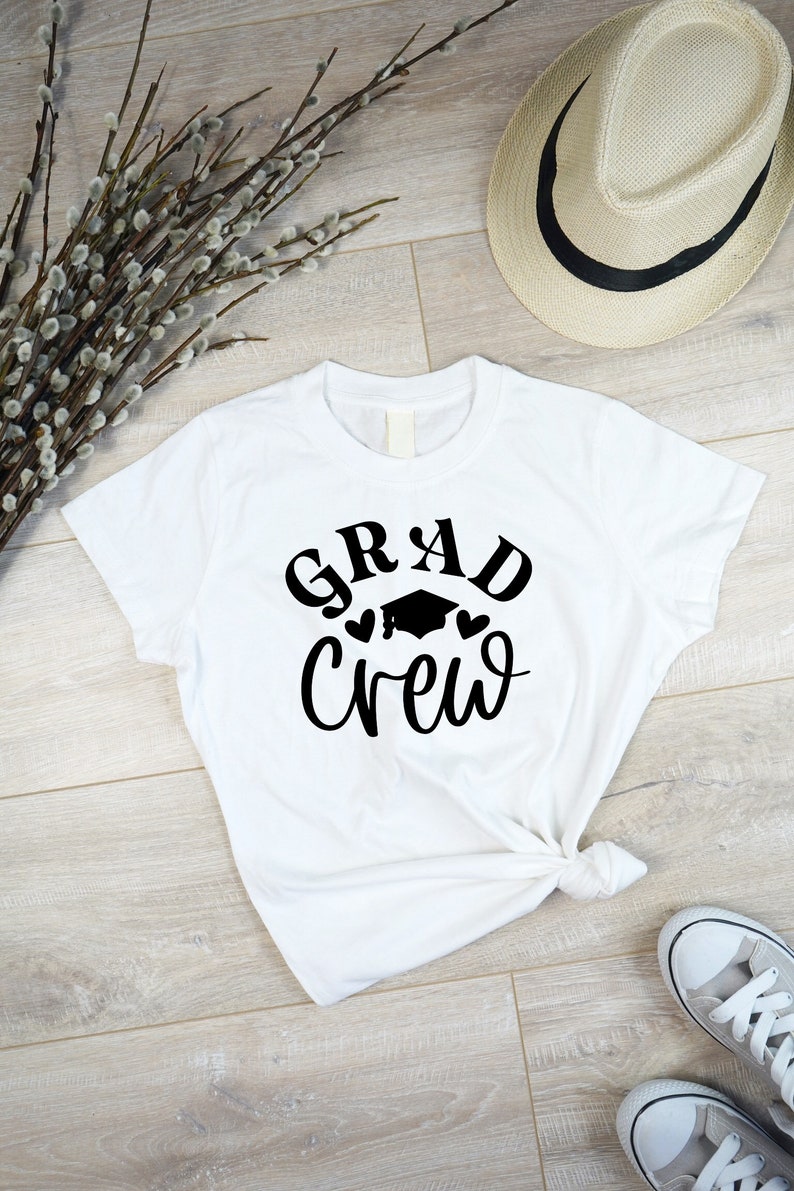 May include: White t-shirt with black text that says "Grad Crew" with a graduation cap and hearts.