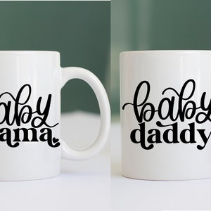 May include: Two white ceramic mugs with black lettering. The mug on the left says "baby mama" and the mug on the right says "baby daddy".