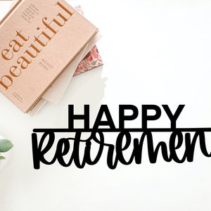 May include: Black metal sign with the text "HAPPY RETIREMENT" in a decorative font. The sign is on a white surface with a book and a potted plant in the background.