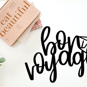 May include: Black and white cursive text that reads "bon voyage" with a small airplane icon next to the word "voyage".