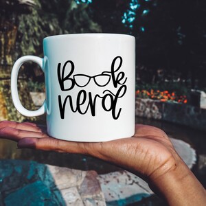 May include: White ceramic mug with a black design that says "Book Nerd" with a pair of glasses above the word "Nerd".