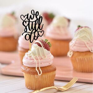May include: A cupcake with pink frosting and a white chocolate-covered strawberry topped with a black cake topper that says "we still do" with hearts.