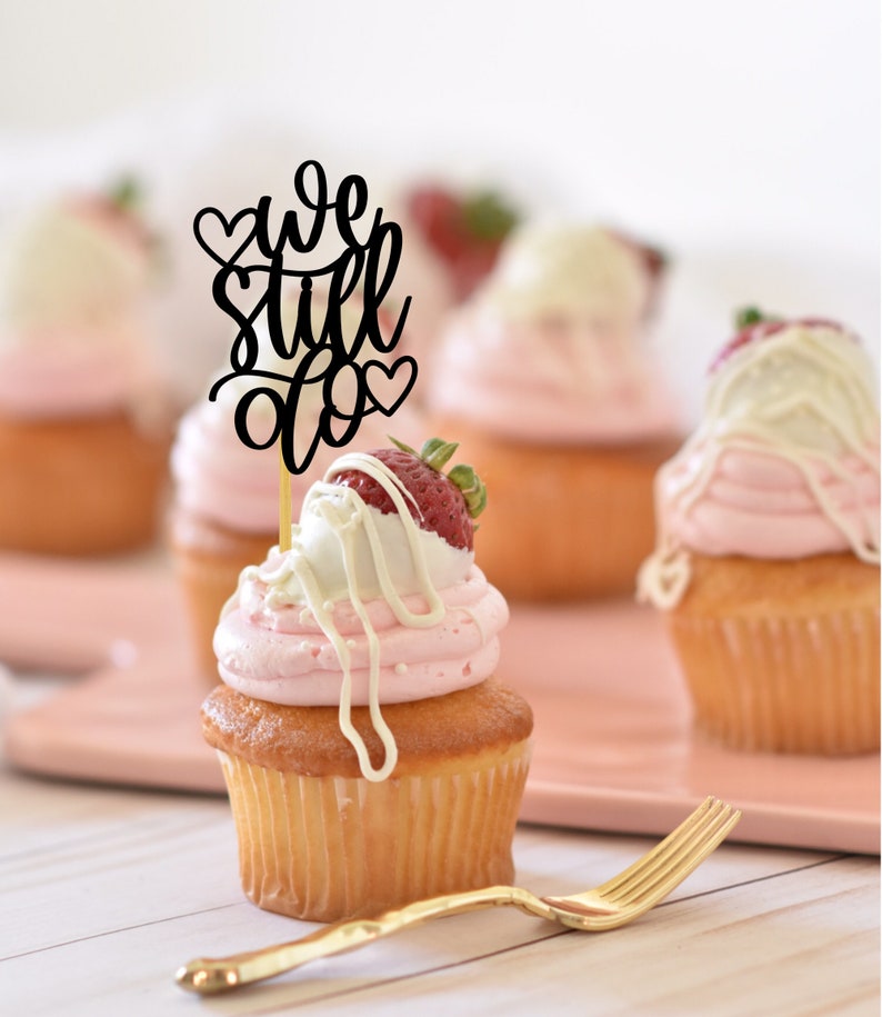 May include: A cupcake with pink frosting and a white chocolate-covered strawberry topped with a black cake topper that says "we still do" with hearts.