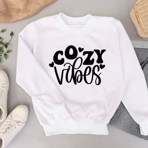 Puede incluir: Sudadera blanca con texto negro que dice "cozy vibes" con corazones alrededor de las letras.