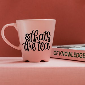 May include: A pink ceramic mug with a black inscription that reads "& that's the tea". The mug is sitting on a pink surface next to a book with the title "OF KNOWLEDGE" printed in green.