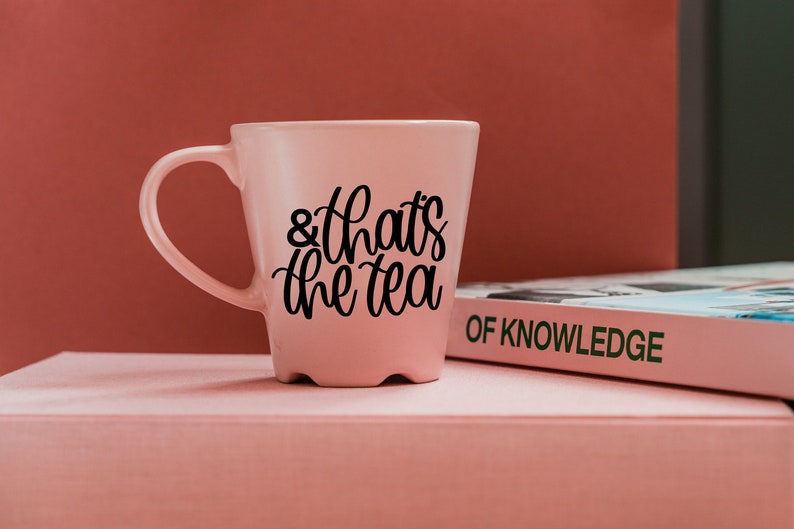 May include: A pink ceramic mug with a black inscription that reads "& that's the tea". The mug is sitting on a pink surface next to a book with the title "OF KNOWLEDGE" printed in green.