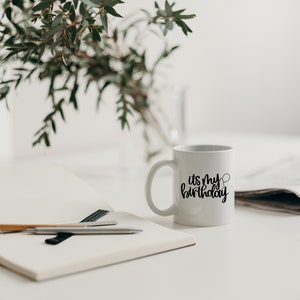 K&ouml;nnte beinhalten: Eine wei&szlig;e Keramiktasse mit schwarzem Text, der "es ist mein Geburtstag" lautet, und einer kleinen Ballongrafik. Die Tasse steht auf einem wei&szlig;en Tisch mit einem Notizbuch, Stiften und einer Zeitung.