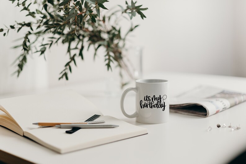 K&ouml;nnte beinhalten: Eine wei&szlig;e Keramiktasse mit schwarzem Text, der "es ist mein Geburtstag" lautet, und einer kleinen Ballongrafik. Die Tasse steht auf einem wei&szlig;en Tisch mit einem Notizbuch, Stiften und einer Zeitung.