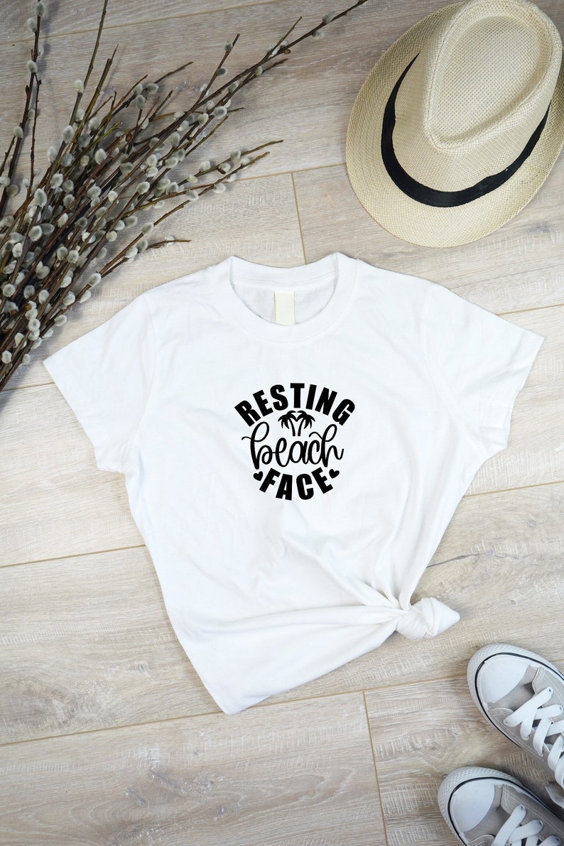 Może przedstawiać: Biała koszulka z czarnym napisem "Resting Beach Face" z palmami i sercami.