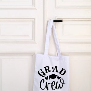 May include: White tote bag with black text that reads "GRAD Crew" with a graduation cap and two hearts.