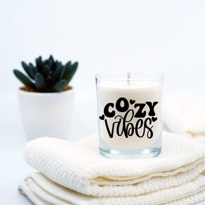 Puede incluir: Una vela blanca en un tarro de vidrio con el texto "cozy vibes" en letras negras. La vela est&aacute; sentada sobre una pila de toallas blancas.