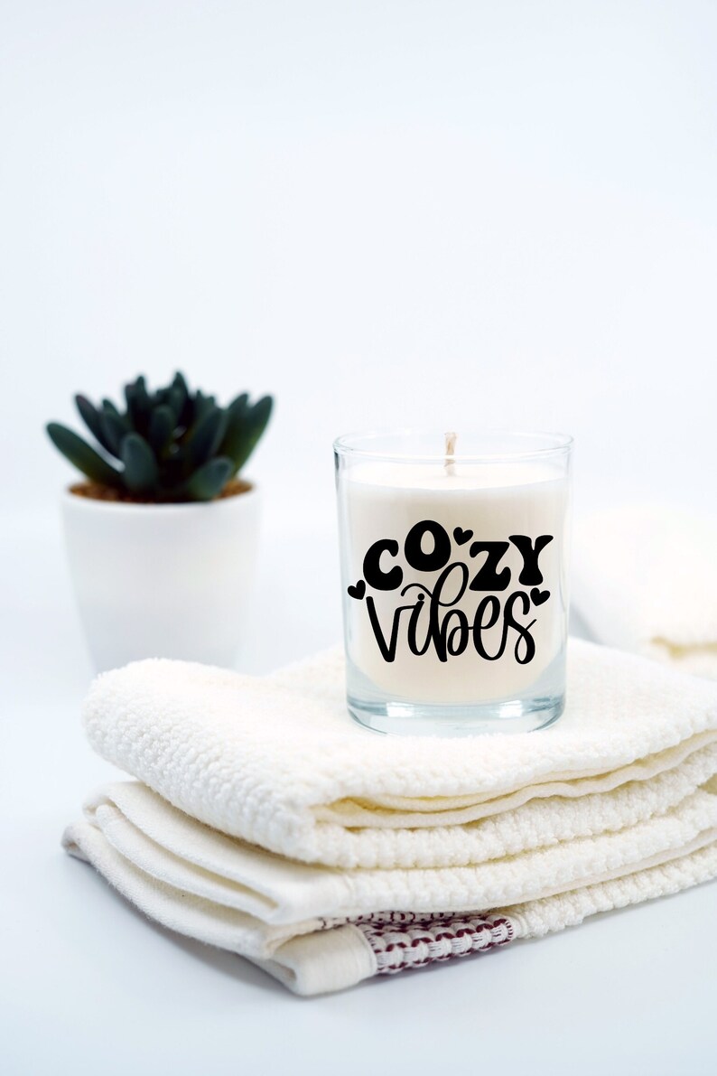 Puede incluir: Una vela blanca en un tarro de vidrio con el texto "cozy vibes" en letras negras. La vela est&aacute; sentada sobre una pila de toallas blancas.