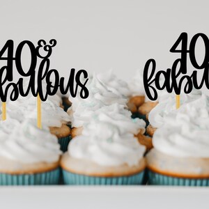 May include: A close-up of cupcakes with black and gold "40 & Fabulous" cake toppers. The cupcakes have white frosting and are arranged in a white tray.