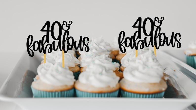 May include: A close-up of cupcakes with black and gold "40 & Fabulous" cake toppers. The cupcakes have white frosting and are arranged in a white tray.
