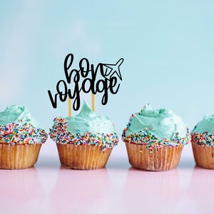 May include: A row of six cupcakes with light blue frosting and colorful sprinkles. The cupcake in the center has a black paper topper that says "bon voyage" with a small airplane graphic.