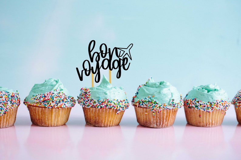 May include: A row of six cupcakes with light blue frosting and colorful sprinkles. The cupcake in the center has a black paper topper that says "bon voyage" with a small airplane graphic.
