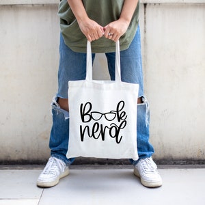 May include: A white tote bag with the text "book nerd" and a pair of glasses printed on it.