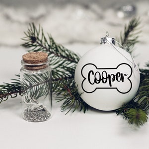 K&ouml;nnte beinhalten: Wei&szlig;er Weihnachtsbaumschmuck in Form eines Knochens mit dem Namen "Cooper" darauf. Der Schmuck ist von gr&uuml;nen Kiefern&auml;sten und einem kleinen Glasgef&auml;&szlig; mit silbernem Glitzer umgeben.