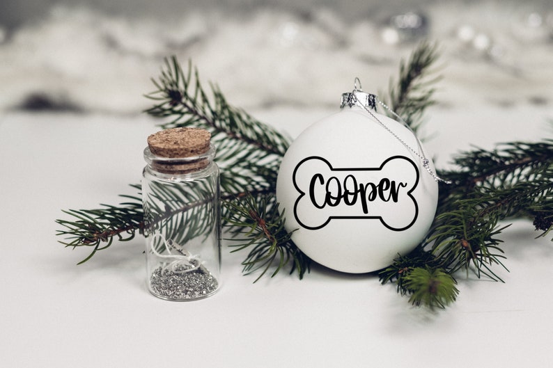 K&ouml;nnte beinhalten: Wei&szlig;er Weihnachtsbaumschmuck in Form eines Knochens mit dem Namen "Cooper" darauf. Der Schmuck ist von gr&uuml;nen Kiefern&auml;sten und einem kleinen Glasgef&auml;&szlig; mit silbernem Glitzer umgeben.