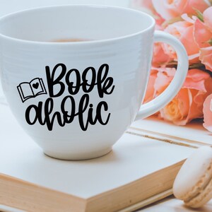 May include: White ceramic mug with a black design that says "Bookaholic" with a small image of an open book and a heart. The mug is sitting on a white surface with a book and a macaron.