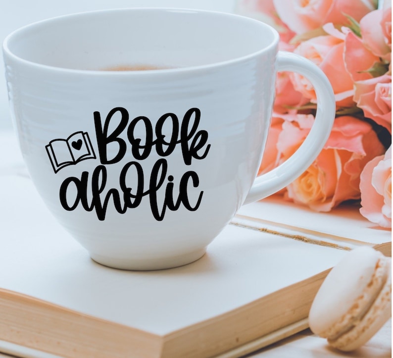 May include: White ceramic mug with a black design that says "Bookaholic" with a small image of an open book and a heart. The mug is sitting on a white surface with a book and a macaron.
