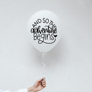 May include: White balloon with black lettering that reads "And so the adventure begins" with a small heart symbol.