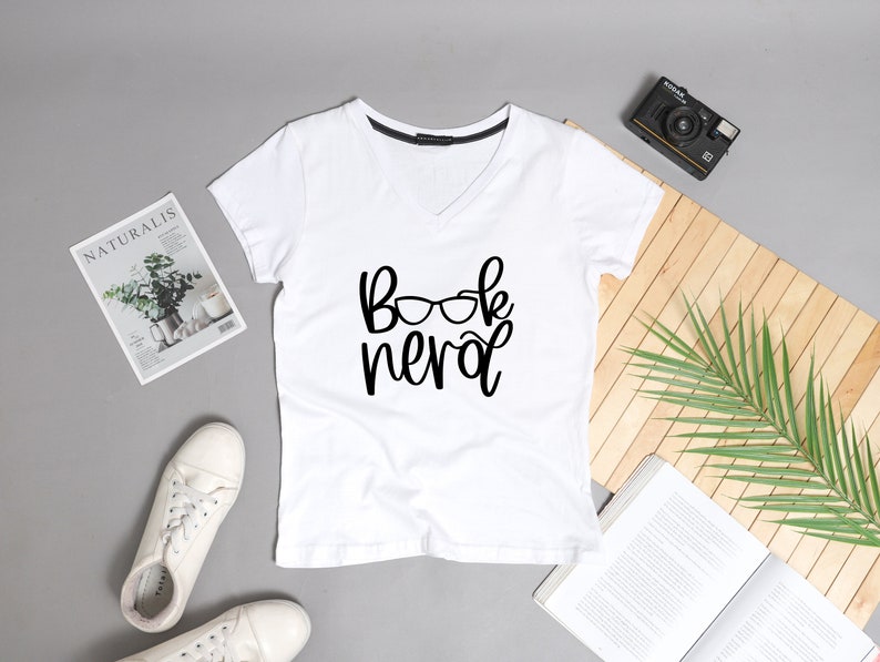 May include: White v-neck t-shirt with the text "Book Nerd" and a pair of glasses graphic printed on the front.