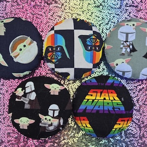 May include: Five circular fabric-covered buttons featuring Star Wars character prints. One button shows Grogu on black, one has a rainbow Darth Vader on a checkerboard, one features Mandalorian characters on gray, one shows Grogu and Mando on black, and one has the Star Wars logo.