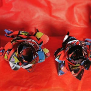May include: Two red, blue, and yellow paper roses made from comic book pages. The roses are on a red background.
